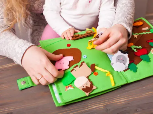 Mani di adulto e bambino esplorano un colorato libro sensoriale in feltro fai da te, con animali e elementi naturali.