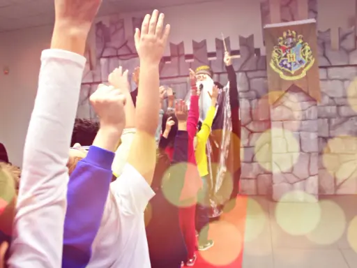 Bambini entusiasti alzano le mani per imparare trucchi di magia facili, con un mago sullo sfondo e uno stemma di Hogwarts.