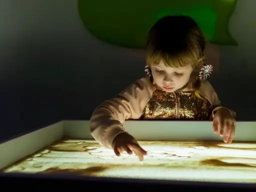 Bambina con i capelli biondi e fiocchi rosa crea disegni nella sabbia illuminata, un momento di sandplay therapy.