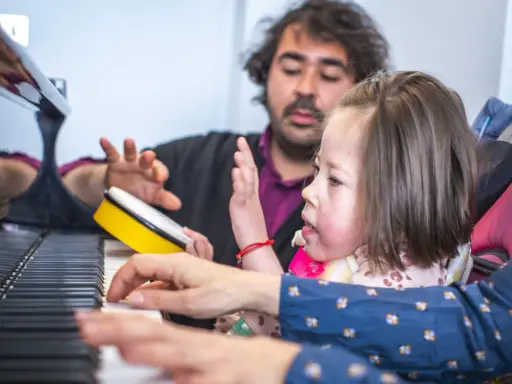 Bambina e terapista suonano il piano, con un tamburello giallo. Esempi di musicoterapia che promuovono l'interazione e l'espressione.