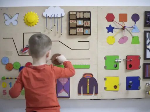Bambino gioca con un pannello sensoriale fai da te: ingranaggi, lucchetti, forme e un gioco del tris.