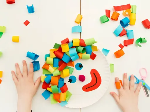 Bambini creano un clown con carta colorata, un'idea per lavoretti di Carnevale. Mani piccole e forbici rosa completano la scena.