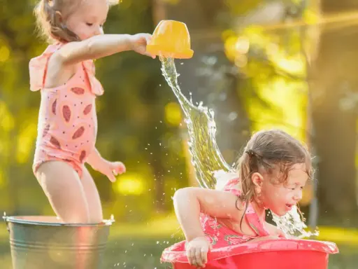 Bambine si divertono con giochi d'acqua al nido, una spruzza l'altra con un secchiello giallo.
