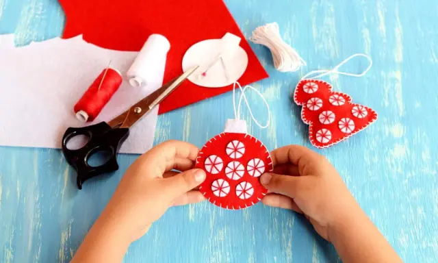 Bambino crea decorazioni natalizie in feltro. Materiali per attività natalizie per bambini: forbici, filo rosso e bianco, feltro rosso e bianco.