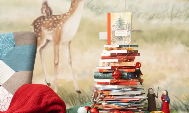 Una pila di libri per le letture di Natale per bambini, decorata con lucine e palline rosse, con un folletto e statuine del presepe.