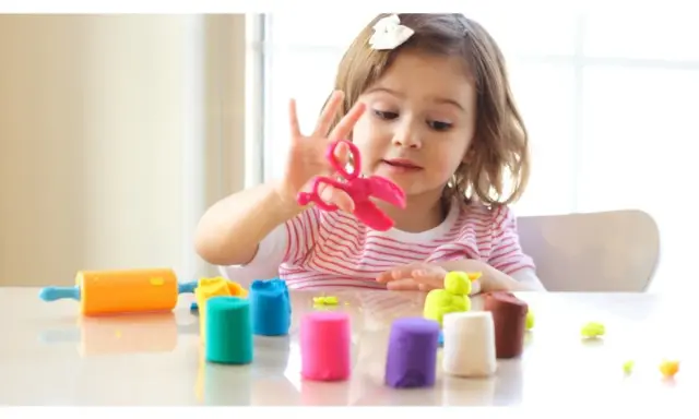 Bambina gioca con pasta modellabile colorata, forbici rosa e mattarello arancione. Giochi di manipolazione Montessori per sviluppare creatività.