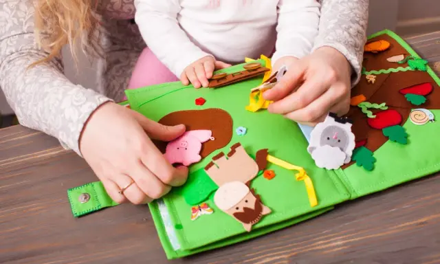 Mani di adulto e bambino esplorano un colorato libro sensoriale in feltro fai da te, con animali e elementi naturali.