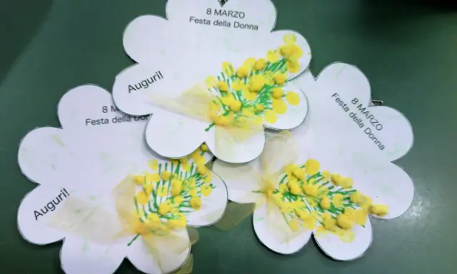 Lavoretto festa della donna infanzia: tre fiori di carta con mimose dipinte e fiocchi gialli. Scritte "Auguri!" e "8 Marzo Festa della Donna".