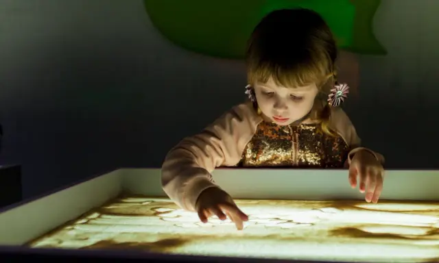 Bambina con i capelli biondi e fiocchi rosa crea disegni nella sabbia illuminata, un momento di sandplay therapy.