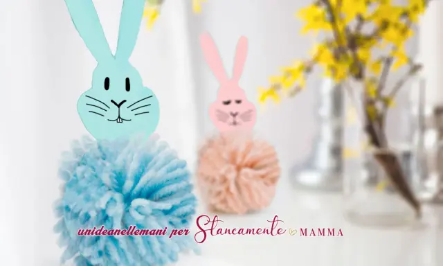Coniglietti con pompon di lana e teste di coniglio origami. Uno è azzurro, l'altro rosa, con fiori gialli sullo sfondo.