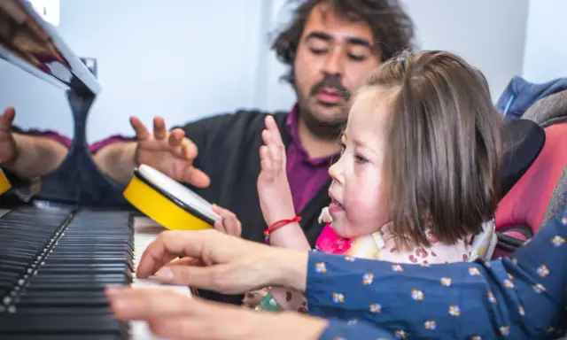 Bambina e terapista suonano il piano, con un tamburello giallo. Esempi di musicoterapia che promuovono l'interazione e l'espressione.