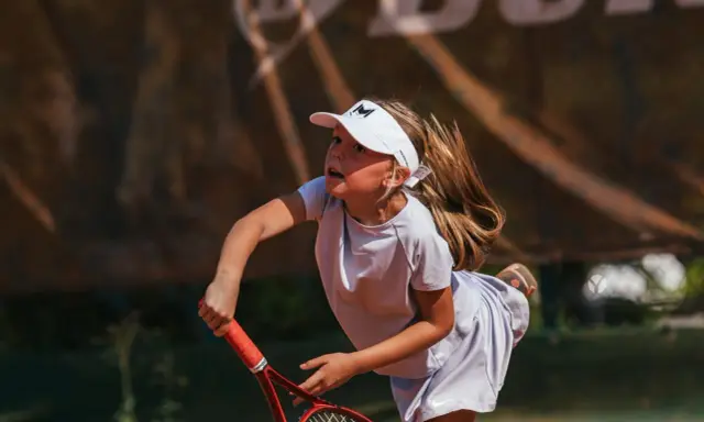 Bambina concentrata durante un servizio di tennis, con la racchetta pronta a colpire la palla.