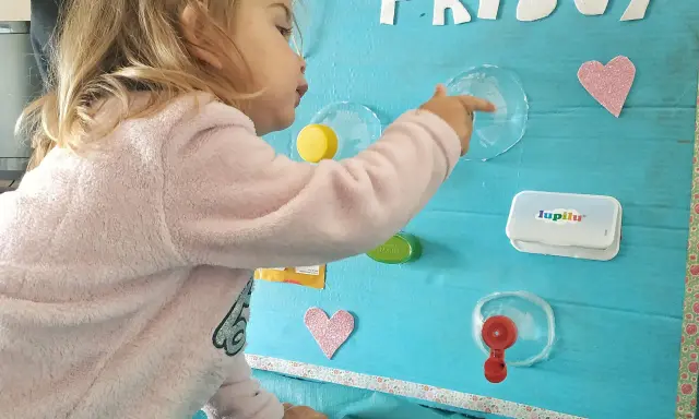 Bambina gioca con giochi motricità fine fai da te: un pannello azzurro con lettere glitterate, cuori rosa e oggetti da manipolare.