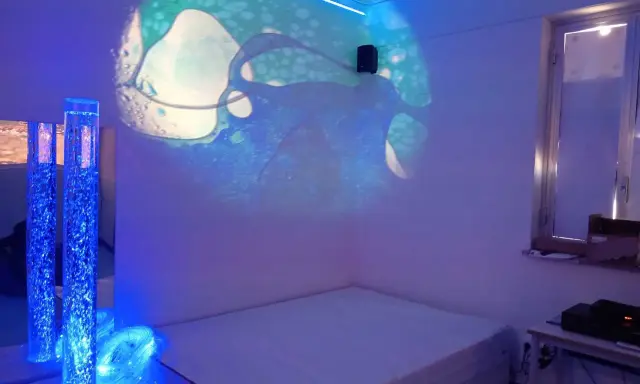 Un'aula snoezelen con proiezioni luminose, colonne d'acqua blu e un letto. Un ambiente rilassante e multisensoriale.