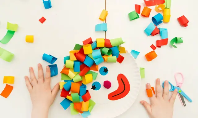 Bambini creano un clown con carta colorata, un'idea per lavoretti di Carnevale. Mani piccole e forbici rosa completano la scena.
