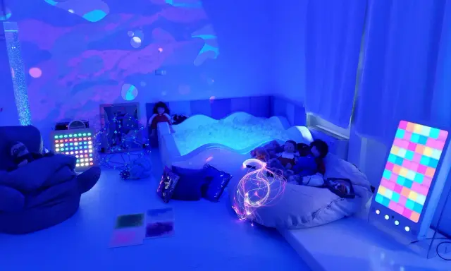 Stanza sensoriale autismo con piscina di palline, luci soffuse, pupazzi e pannelli interattivi per stimolare i sensi.