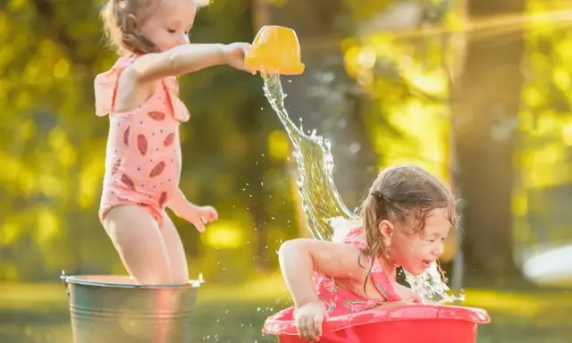 Bambine si divertono con giochi d'acqua al nido, una spruzza l'altra con un secchiello giallo.