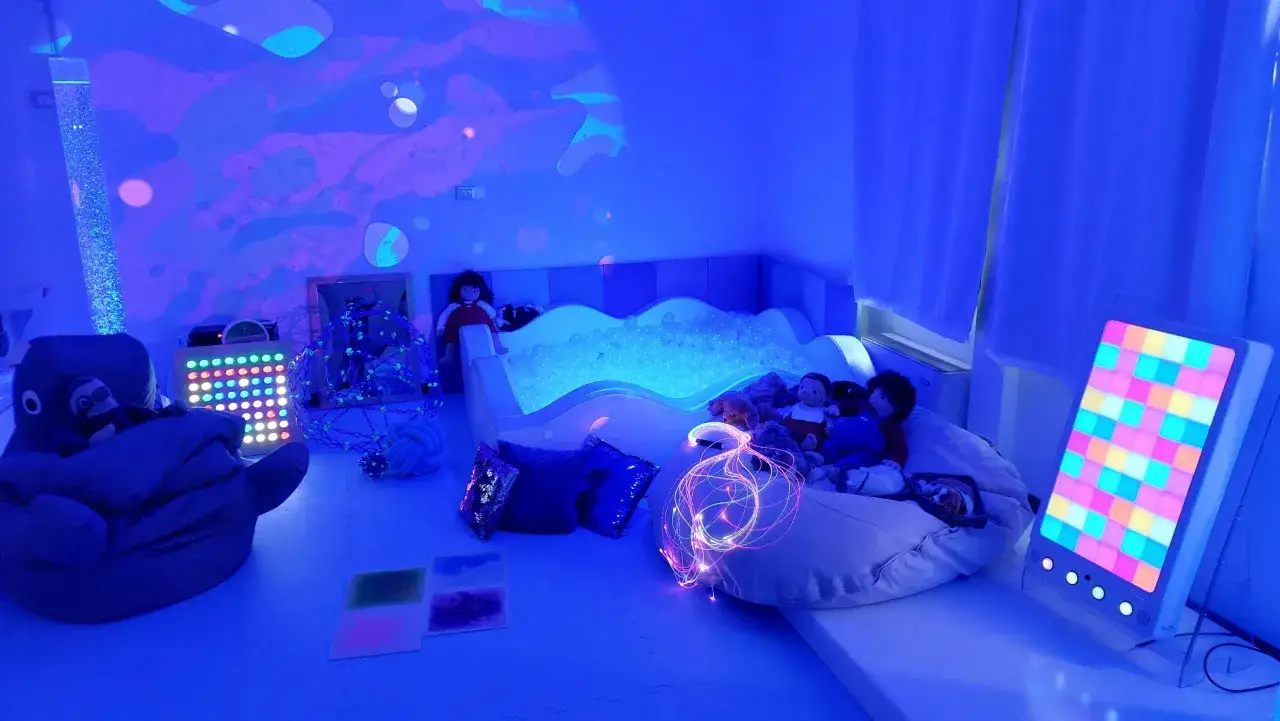 Un'aula snoezelen con luci soffuse, una piscina di palline, pupazzi e pannelli interattivi per stimolare i sensi.