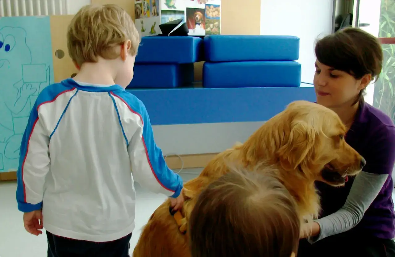 Bambini e cane in un momento di pet therapy. Un bimbo accarezza un golden retriever mentre una donna lo osserva sorridendo.