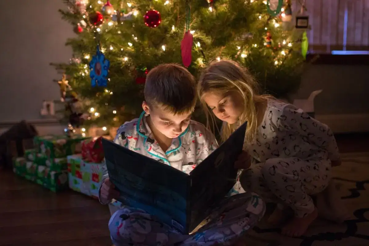 Bambini in pigiama leggono un libro sotto l'albero di Natale illuminato, creando una magica storia sul natale.