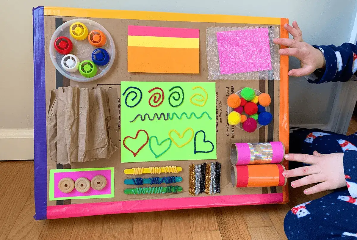 Pannello con giochi sensoriali tattili: pompon, fili colorati, carta da pacchi, bolle di plastica e rotoli decorati.