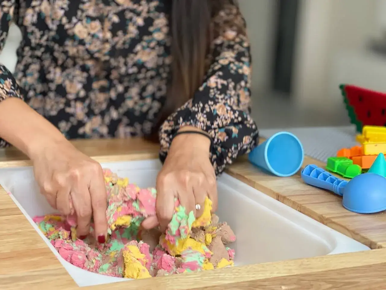 Mani che manipolano sabbia cinetica colorata, ideale per giocare da piccolissimi. Accessori da spiaggia e blocchi colorati sullo sfondo.