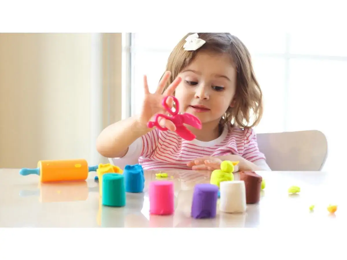 Bambina gioca con pasta modellabile colorata, forbici rosa e mattarello arancione. Giochi di manipolazione Montessori per sviluppare creativit&agrave; e manualit&agrave;.