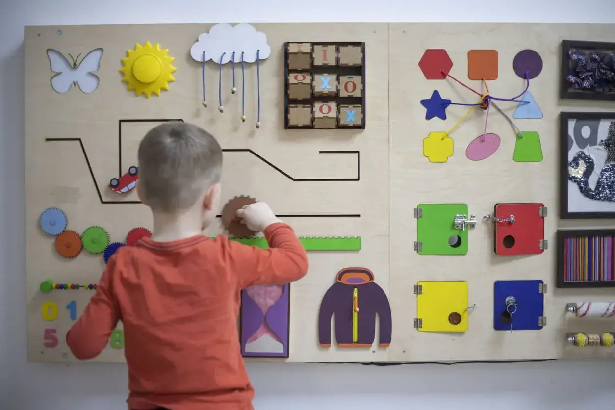 Bambino gioca con un pannello sensoriale fai da te: ingranaggi, lucchetti, forme e un gioco del tris.