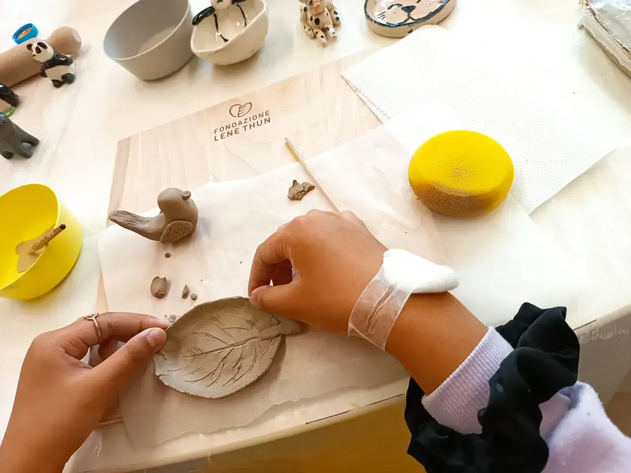 Mani che lavorano la creta per creare una foglia e un uccellino. Attrezzi e materiali sono sparsi sul tavolo.