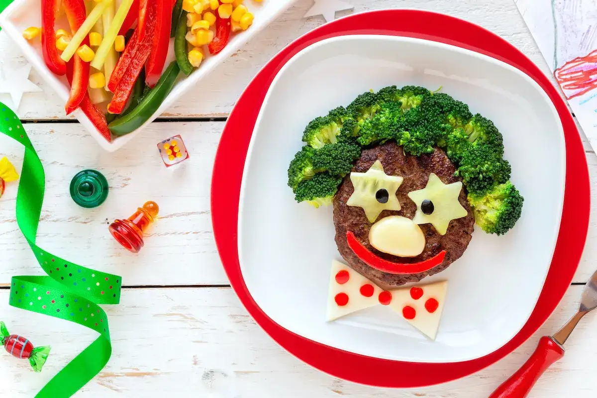 Un piatto divertente con un clown fatto di cibo, perfetto per i giochi sull'alimentazione per bambini. Broccoli per capelli, formaggio a stella per occhi, un naso di formaggio e un sorriso di peperone rosso.