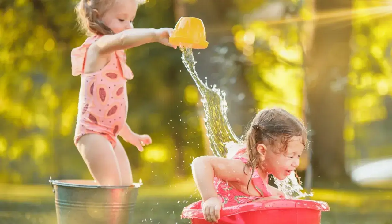 Bambine si divertono con giochi d'acqua al nido, una spruzza l'altra con un secchiello giallo.