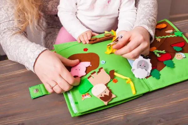 Mani di adulto e bambino esplorano un colorato libro sensoriale in feltro fai da te, con animali e elementi naturali.