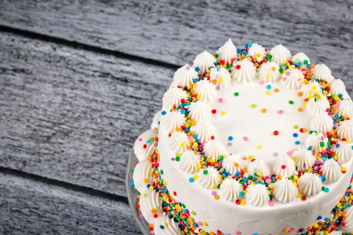 Torta compleanno bianca con glassa, decorata con ciuffi di panna e confettini colorati.