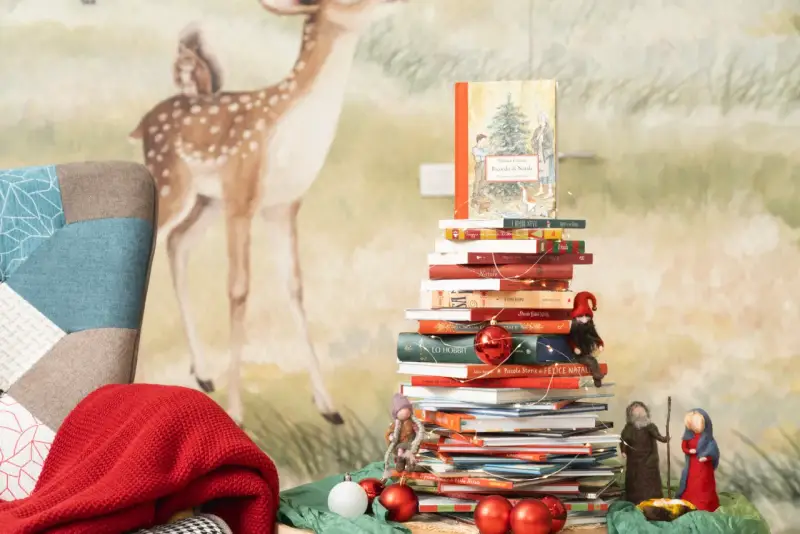 Una pila di libri per le letture di Natale per bambini, decorata con lucine e palline rosse, con un folletto e statuine del presepe.
