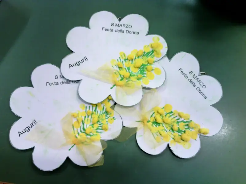 Lavoretto festa della donna infanzia: tre fiori di carta con mimose dipinte e fiocchi gialli. Scritte "Auguri!" e "8 Marzo Festa della Donna".