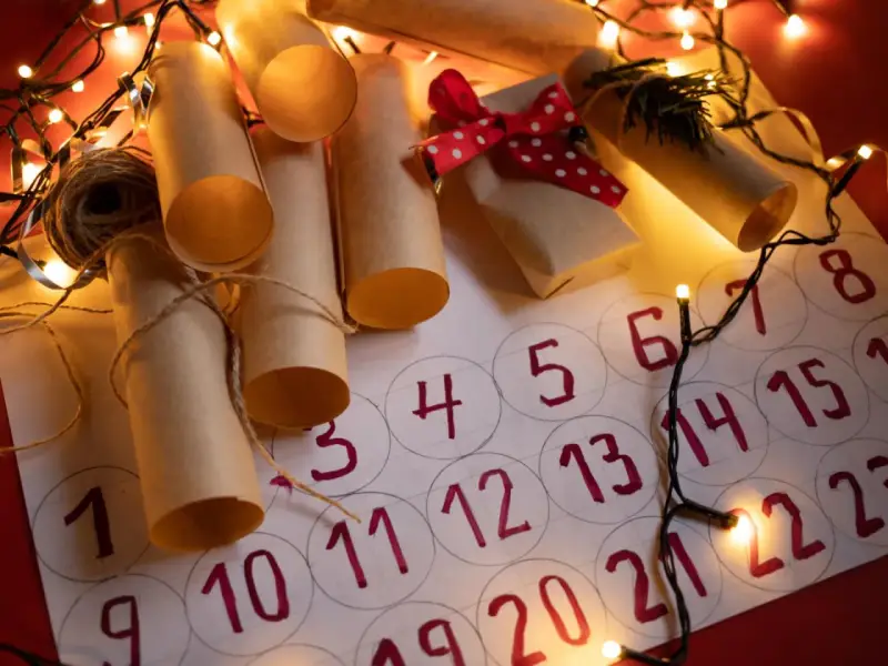 Un **calendario avvento fai da te cartoncino** con rotoli di carta marrone legati con spago, luci natalizie e un piccolo pacchetto regalo.